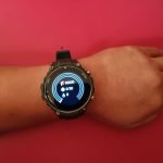 Smartwatch Q70 Pro, en la muñeca, de color verde y de fondo rojo, mostrando sus funcionalidades