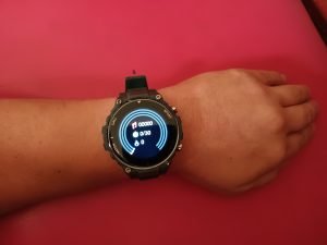 Smartwatch Q70 Pro, en la muñeca, de color verde y de fondo rojo, mostrando sus funcionalidades